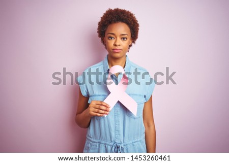 Young african american woman holding brest cancer ribbon over isolated pink background with a confident expression on smart face thinking serious