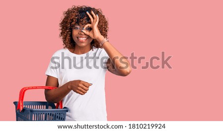Young african american woman holding supermarket shopping basket smiling happy doing ok sign with hand on eye looking through fingers 