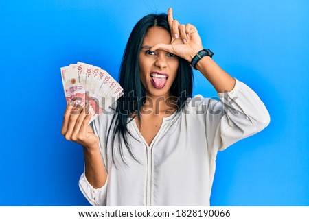 Young african american woman holding 10 colombian pesos banknotes making fun of people with fingers on forehead doing loser gesture mocking and insulting. 