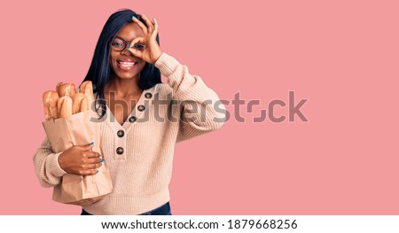Young african american woman holding paper bag with bread smiling happy doing ok sign with hand on eye looking through fingers 