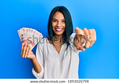 Young african american woman holding 10 colombian pesos banknotes pointing to you and the camera with fingers, smiling positive and cheerful 
