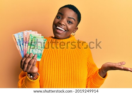 Young african american woman holding south african rand banknotes celebrating achievement with happy smile and winner expression with raised hand 