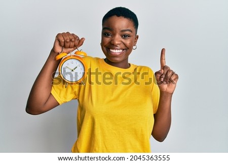 Young african american woman holding alarm clock smiling with an idea or question pointing finger with happy face, number one 