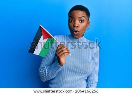 Young african american woman holding palestine flag scared and amazed with open mouth for surprise, disbelief face 