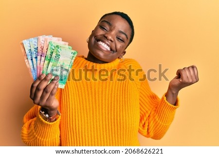 Young african american woman holding south african rand banknotes screaming proud, celebrating victory and success very excited with raised arm 