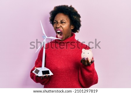 Young african american woman holding windmill and piggy bank angry and mad screaming frustrated and furious, shouting with anger. rage and aggressive concept. 