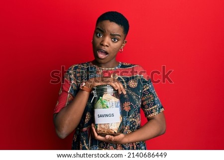 Young african american woman holding savings jar with south african rands money in shock face, looking skeptical and sarcastic, surprised with open mouth 