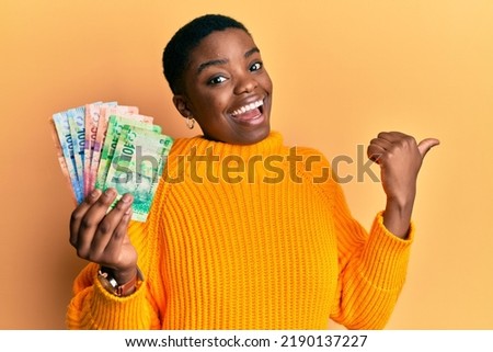Young african american woman holding south african rand banknotes pointing thumb up to the side smiling happy with open mouth 
