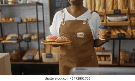 Young african american woman holds coffee and pastries in a bakery shop interior, showcasing various breads and baked goods with wooden shelves in the background, wearing a brown apron - Powered by Shutterstock