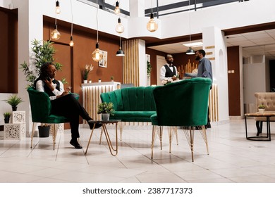 Young African American woman hotel front desk receptionist talking on phone and taking notes while sitting on lobby. Female hospitality manager making phone call ordering supplies - Powered by Shutterstock