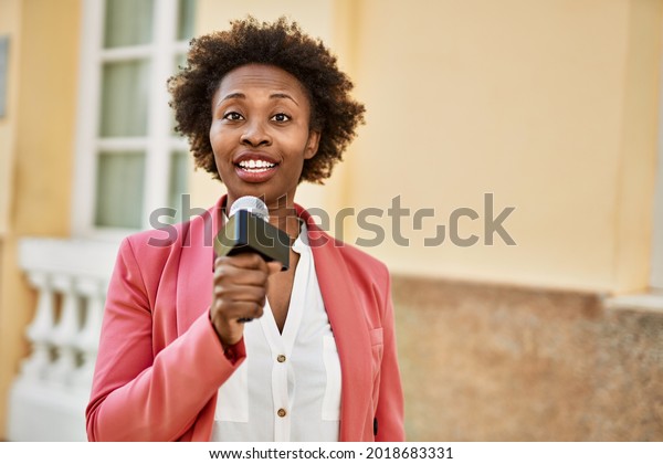 Young african american woman journalist holding\
reporter microphone speaking and smiling to the camera for\
television news
