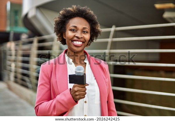 Young african american woman journalist holding
reporter microphone speaking and smiling to the camera for
television news