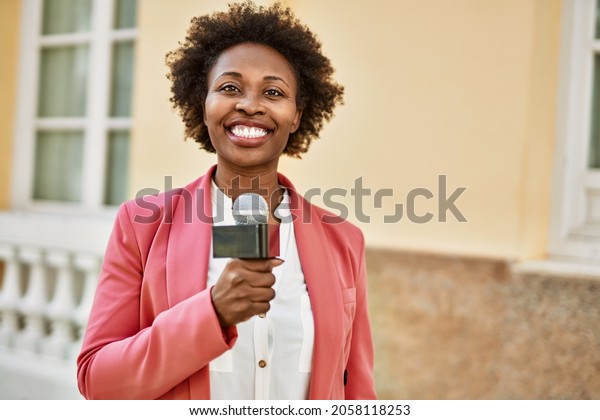 Young african american woman journalist holding\
reporter microphone speaking and smiling to the camera for\
television news