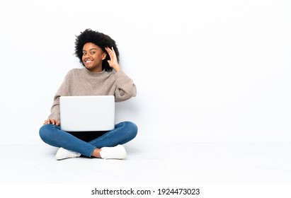 Young African American woman with a laptop sitting on the floor listening to something by putting hand on the ear