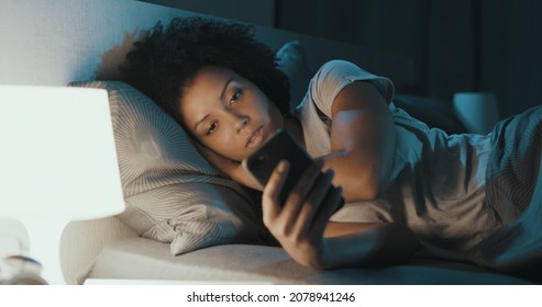 Young African American Woman Lying In Bed At Night, She Can't Sleep And She Is Checking Her Smartphone