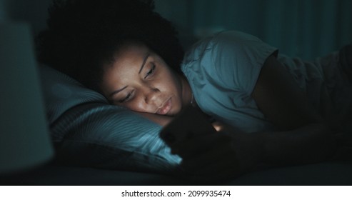 Young African American Woman Lying In Bed At Night, She Can't Sleep And She Is Checking Her Smartphone