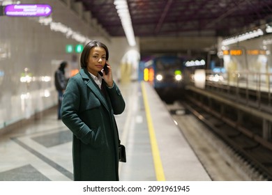 Young african american woman office worker making phonecall while standing at subway platform after workday and waiting for train. African female in coat talking by mobile phone at underground station - Shutterstock ID 2091961945