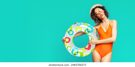 A young African American woman in an orange swimsuit stands against a turquoise background, holding a colorful inflatable ring. She is wearing a straw hat and sunglasses, copy space - Powered by Shutterstock