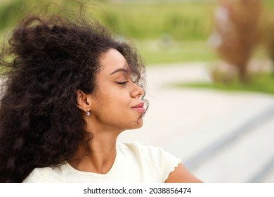 Young African American Woman Outdoors On Nature Background. Attractive Mixed Race Girl With Wind In Loose Wavy Curls African American Hair And Closed Eyes Enjoying Fresh Air. Soft Focus. Side View