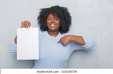 Young African American Woman Over Grey Grunge Wall Holding Blank Paper Sheet With Surprise Face Pointing Finger To Herself