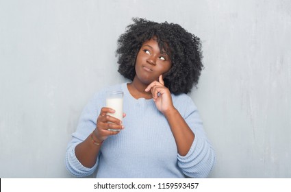 Young african american woman over grey grunge wall drinking a glass of milk serious face thinking about question, very confused idea