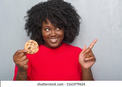 Young african american woman over grey grunge wall eating chocolate chip cooky very happy pointing with hand and finger to the side