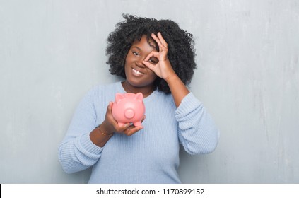 Young african american woman over grey grunge wall holding piggy bank with happy face smiling doing ok sign with hand on eye looking through fingers Stock Photo