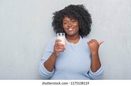 Young african american woman over grey grunge wall drinking a glass of milk pointing and showing with thumb up to the side with happy face smiling