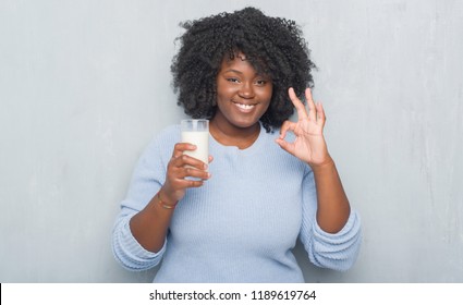 Young african american woman over grey grunge wall drinking a glass of milk doing ok sign with fingers, excellent symbol