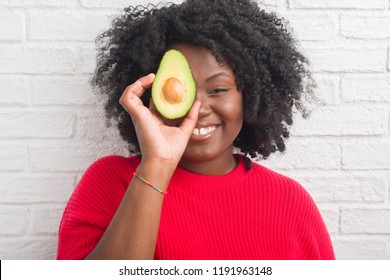 Young african american woman over white brick wall eating avocado with a happy face standing and smiling with a confident smile showing teeth