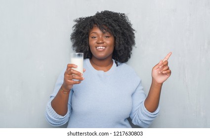 Young african american woman over grey grunge wall drinking a glass of milk very happy pointing with hand and finger to the side