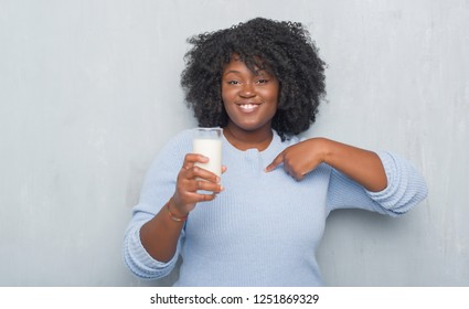 Young african american woman over grey grunge wall drinking a glass of milk with surprise face pointing finger to himself