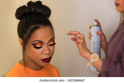 Young African American Woman  With Perfect Skin Facial. Fashion Luxury Makeup Orange-coloured Shades. Long Eyelashes, Pouty Lips. Upper Bun Hair. Professional Hair Stylist Uses Hairspray To Fix Hair.