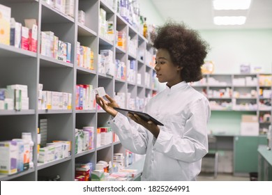 Young African American Woman Pharmacist Or Chemist Using Digital Tablet, While Standing Near The Shelves With Medicines In Modern Pharmacy During Drugs Inventory