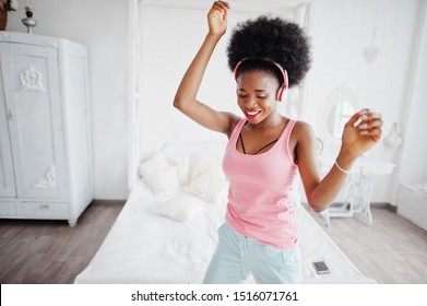 Young African American Woman In Pink Singlet Dancing And Listen Music On Earphones At Her White Room.