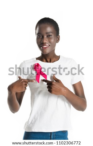 young african american woman pointing at pink ribbon and smiling at camera isolated on white, breast cancer concept