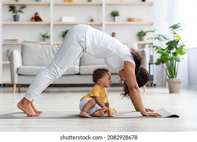Young African American Woman Practicing Yoga With Her Little Baby Son At Home, Sporty Black Female Making Back Stretching Exercise, Standing In Dog Pose While Training On Fitness Mat In Living Room