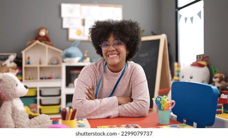 Young african american woman preschool teacher smiling confident sitting on table with arms crossed gesture at kindergarten - Shutterstock ID 2299079737
