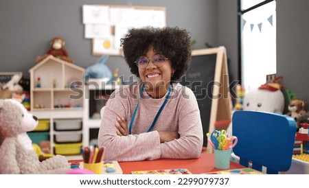 Young african american woman preschool teacher smiling confident sitting on table with arms crossed gesture at kindergarten
