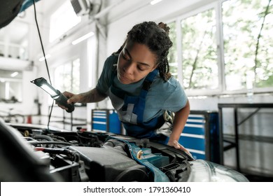 Young african american woman, professional female mechanic examining under hood of car with torch at auto repair shop. Car service, maintenance and people concept. Selective focus. Horizontal shot - Powered by Shutterstock