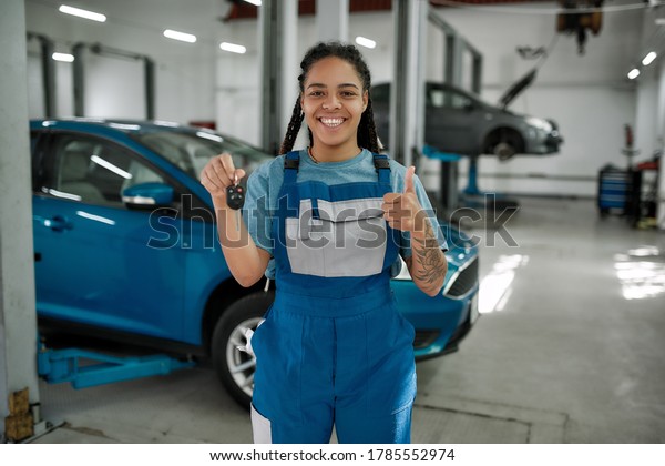 Young african american woman, professional female\
mechanic smiling at camera, giving car key and showing thumbs up in\
auto repair shop. Car service concept. Focus on person. Horizontal\
shot