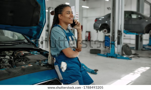 Young african american woman, professional female\
mechanic talking on phone, leaning on a car with open hood at auto\
repair shop. Car service, maintenance and people concept. Side\
view. Web Banner