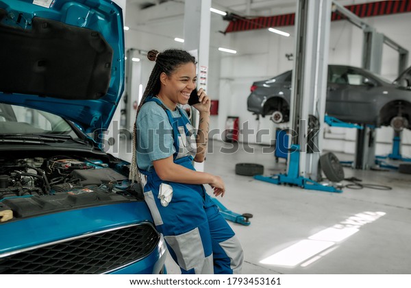 Young african american woman, professional female\
mechanic talking on phone, leaning on car with open hood at auto\
repair shop. Car service, maintenance and people concept. Side\
view. Horizontal shot