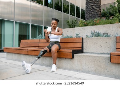 Young African American woman with a prosthetic leg, wearing a tank top and camo shorts, sits on a bench. The woman with a prosthetic leg checks her smartwatch. Inclusive sports with disabled athlete. - Powered by Shutterstock