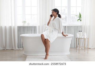 A young African American woman relaxes in her bright bathroom, talking on her phone while pampering herself in a cozy robe. Sunlight filters through sheer curtains, enhancing her peaceful moment. - Powered by Shutterstock