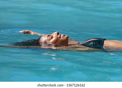 Young african american woman relaxing in swimming pool at spa resort. Portrait of beautiful black girl in bikini floating on water. Side view of cheerful tanned woman enjoying summer vacation.