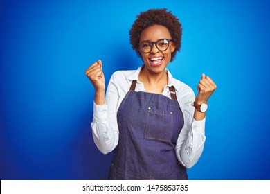 Young African American Woman Shop Owner Wearing Business Apron Over Blue Background Celebrating Surprised And Amazed For Success With Arms Raised And Open Eyes. Winner Concept.