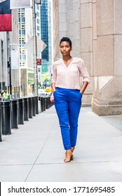 Young African American Woman With Short Afro Hair Traveling In New York City, Wearing Light Color V Neck Shirt, Blue Pants, Brown Sandal High Heels, Walking On Narrow Old Street With High Buildings.