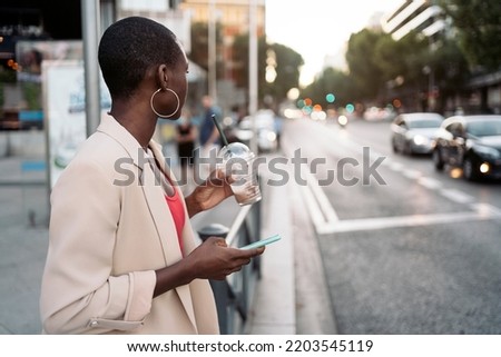 Young african american woman with short hair and suit holding phone and a milkshake waiting and looking to the side to cross the street