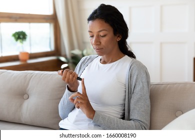 Young African American woman sit on couch at home do daily checkup glucose test with glucometer, biracial millennial female pinch finger measure blood sugar level at home, diabetes problem concept